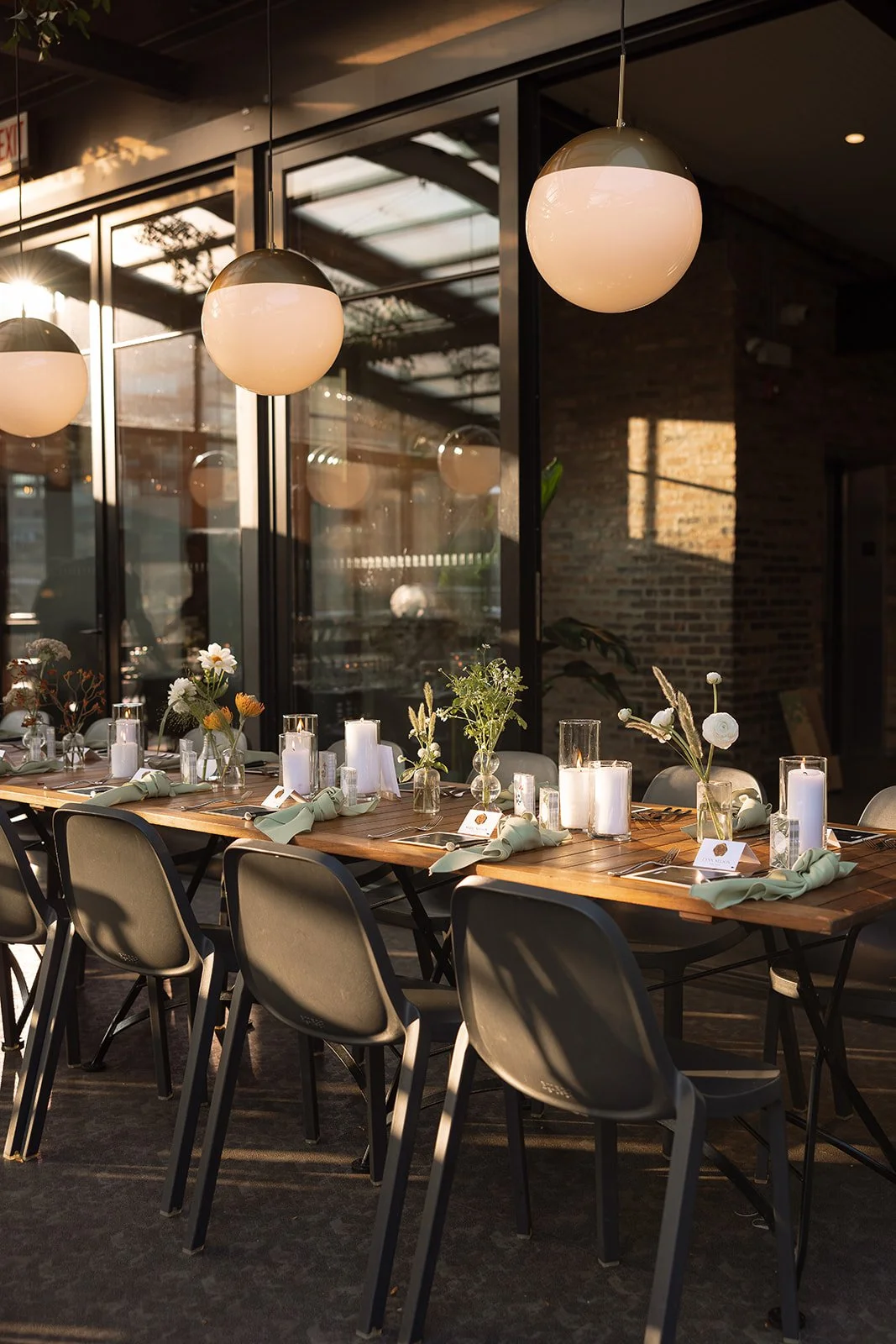 Elegant dining table setup with chairs, vases with flowers, candles, and napkins, surrounded by large hanging spherical lights in a modern dining area.