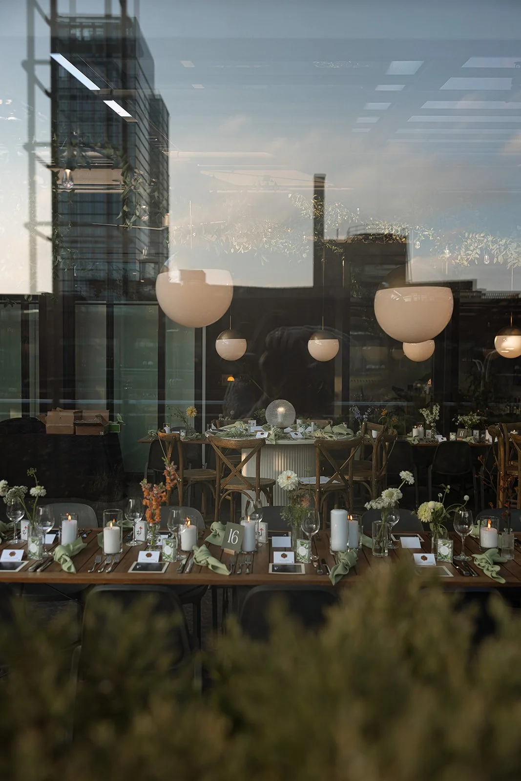 Reflection of a city building in a window showing an elegant indoor dining setup with wooden tables, chairs, and decorative table settings including candles, flowers, and place cards.