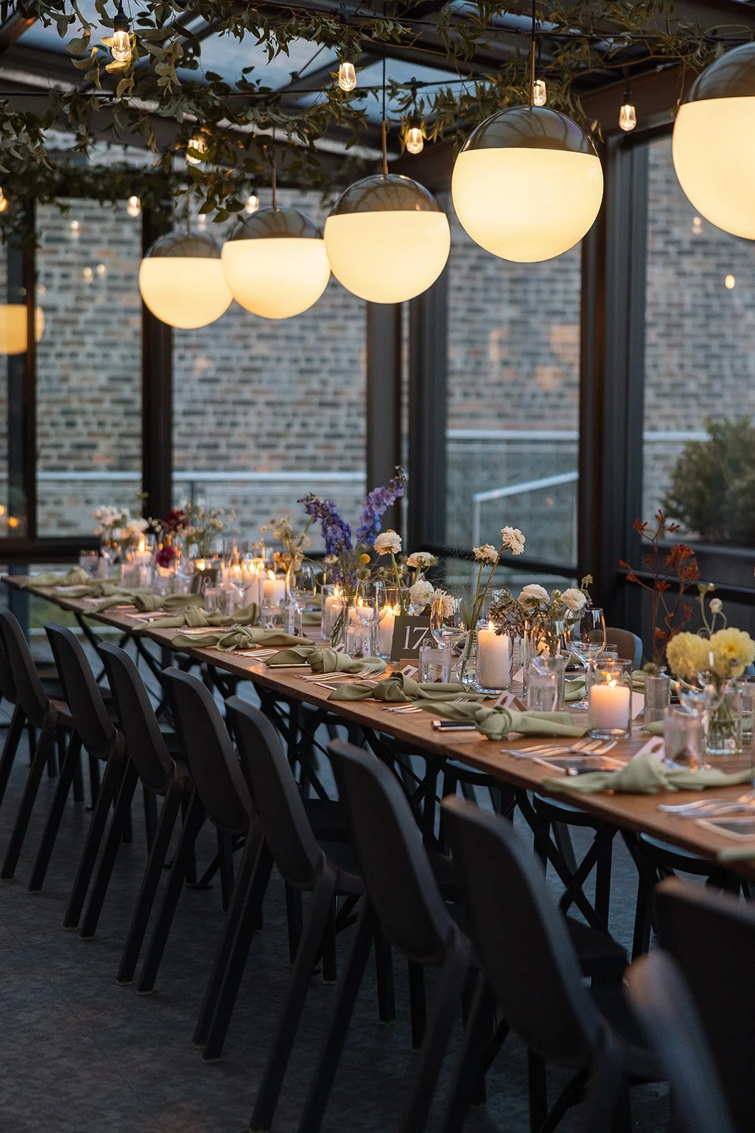 Elegant indoor dining setup with a long table, black chairs, and floral centerpieces under round pendant lights and greenery.