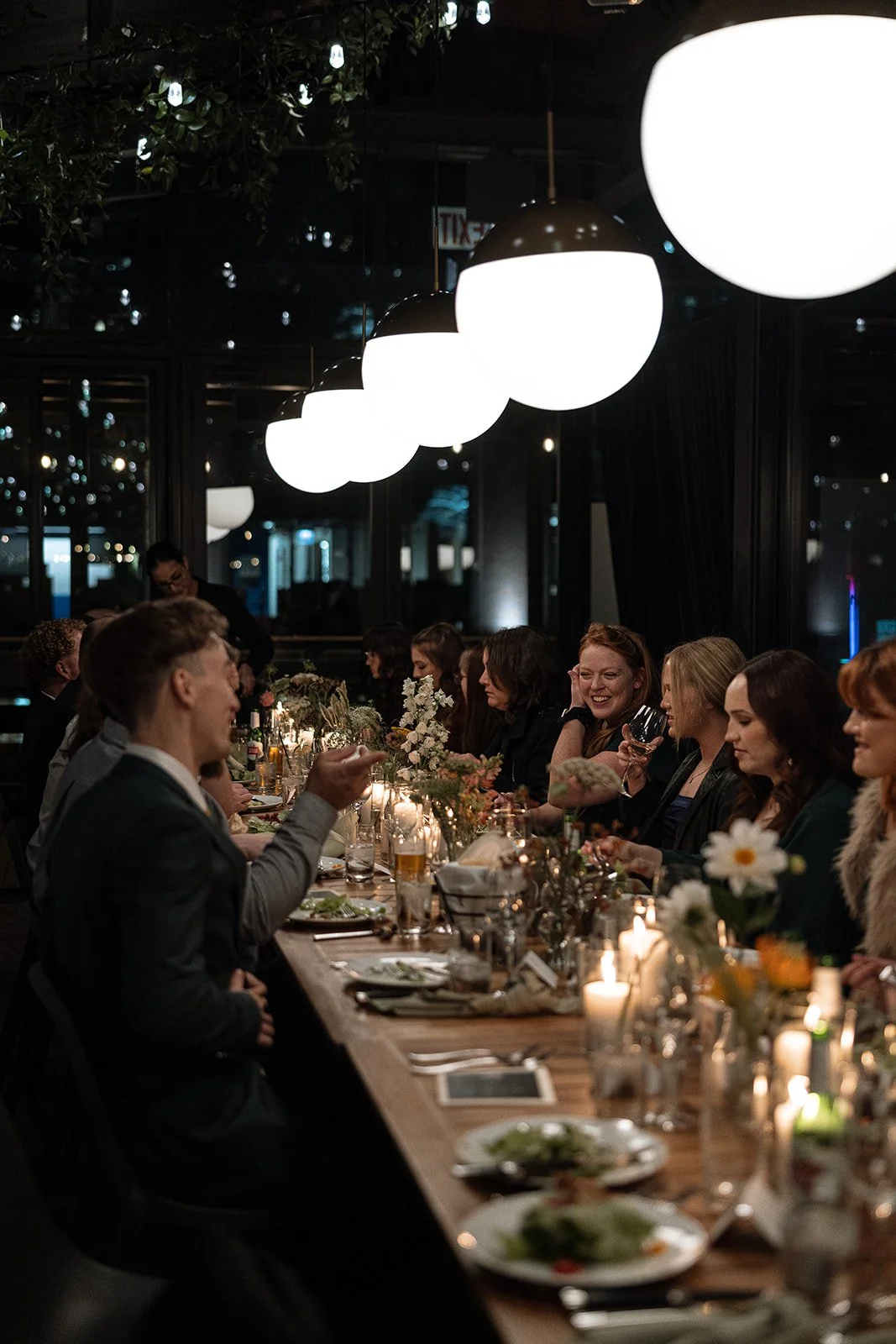 Group of people dining at a long wooden table in a dimly lit restaurant with large round lights overhead and decorative flowers and candles.