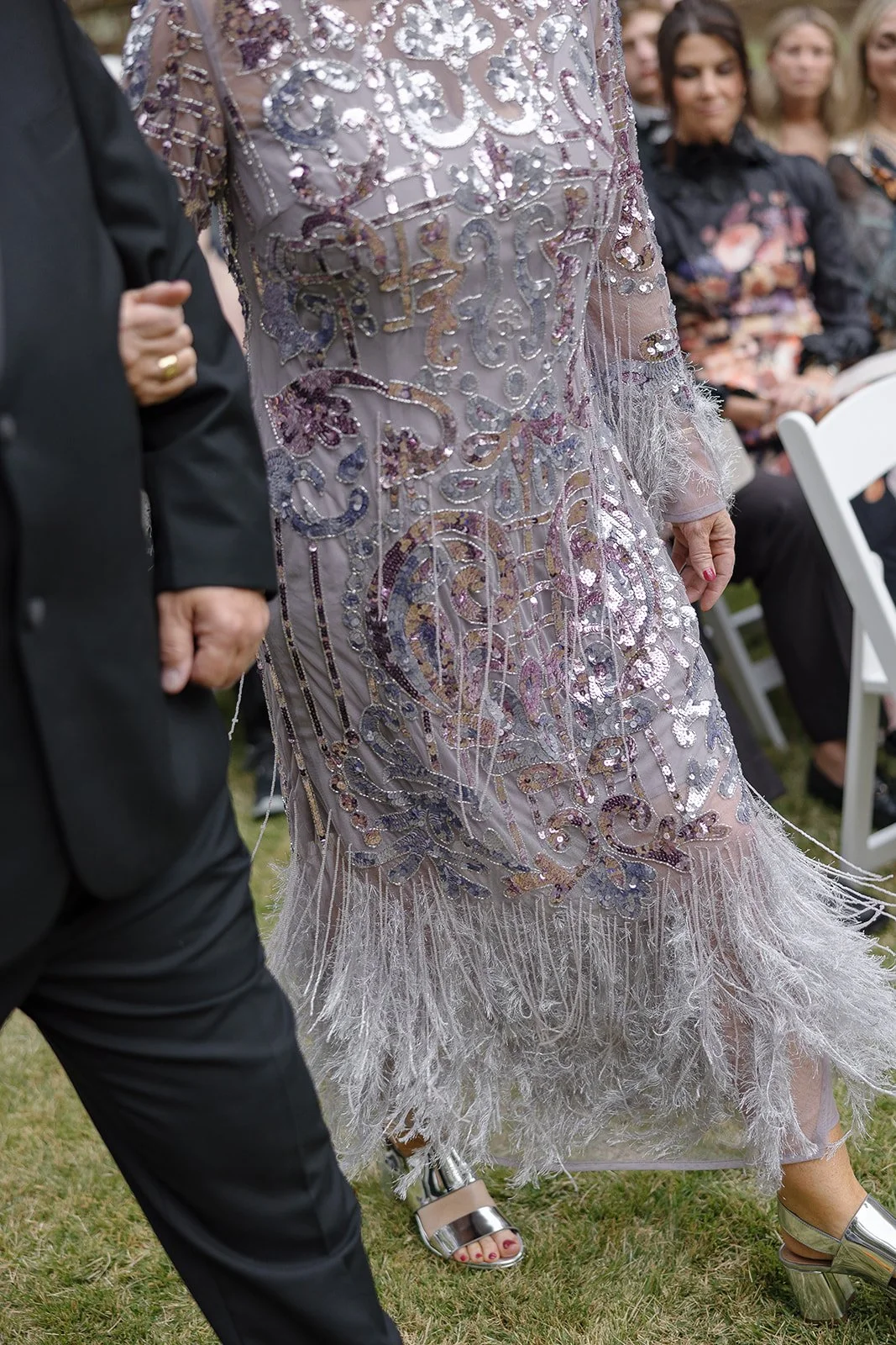 Person in a sequined dress walking, holding arm of another in a black suit at an outdoor event.