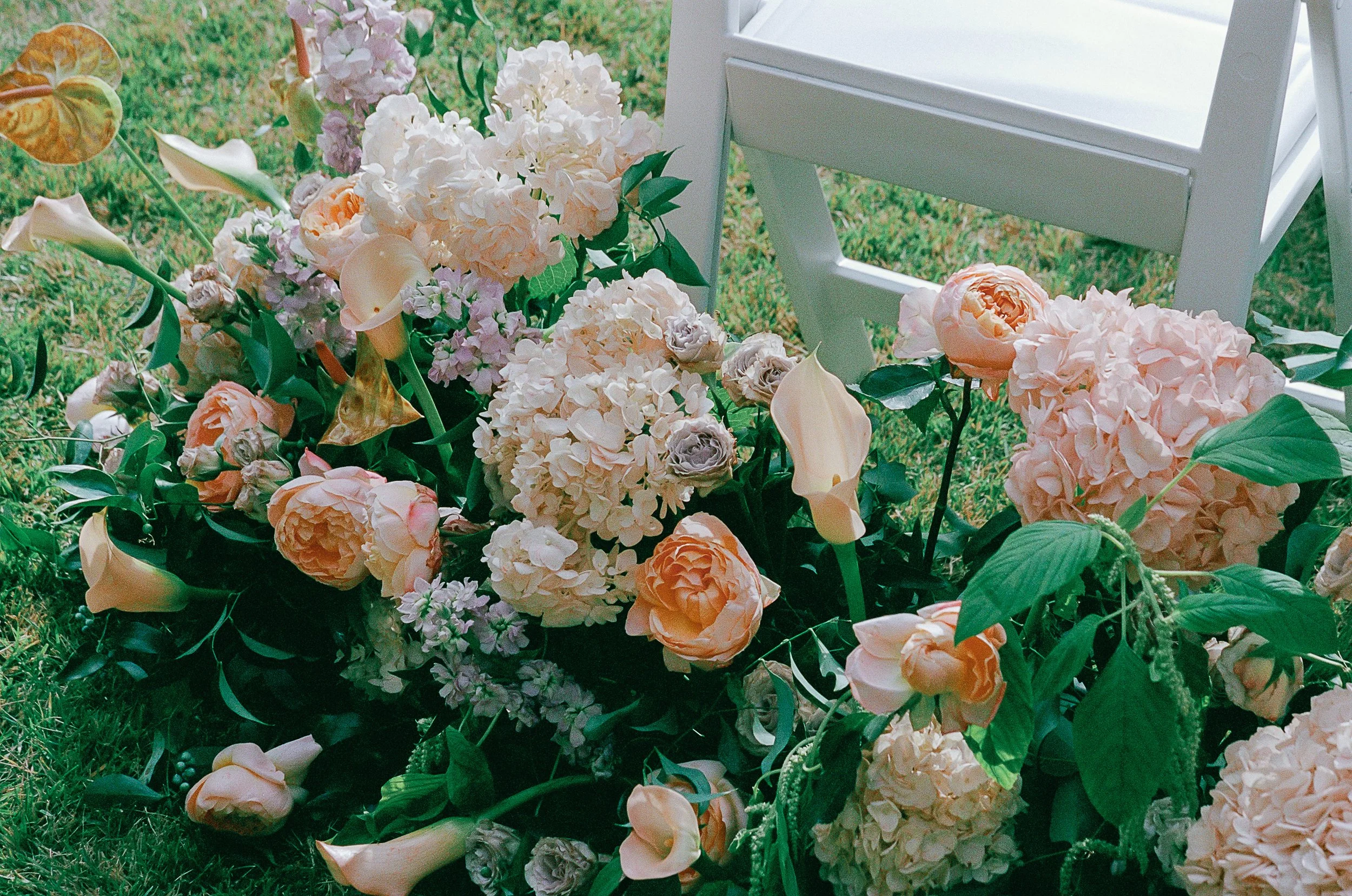 Floral arrangement with roses, hydrangeas, calla lilies, and other flowers next to a white folding chair on grass.