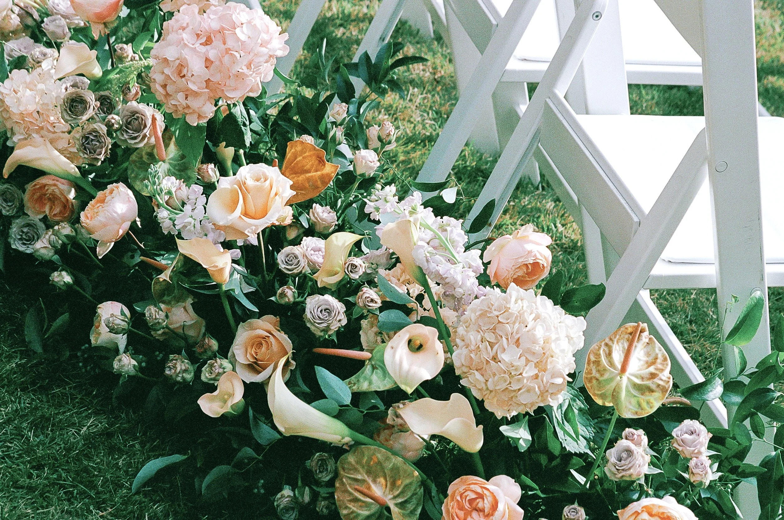 Floral arrangement with roses, peonies, hydrangeas, and greenery beside white folding chairs on grass.