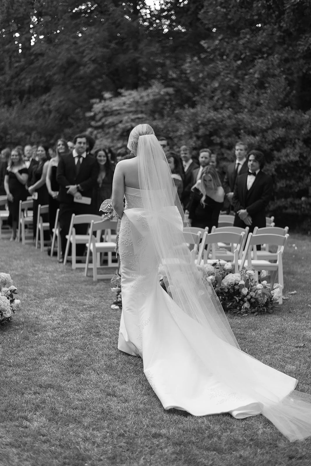 Bride in white dress and veil walking down outdoor aisle, surrounded by guests seated on white chairs.