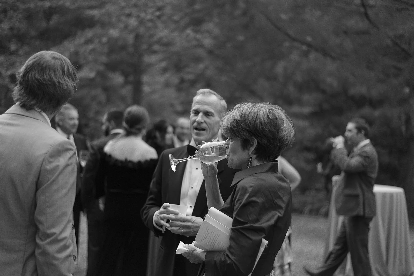 Black and white image of people in formal attire at an outdoor social event, with a woman drinking from a glass in the foreground.