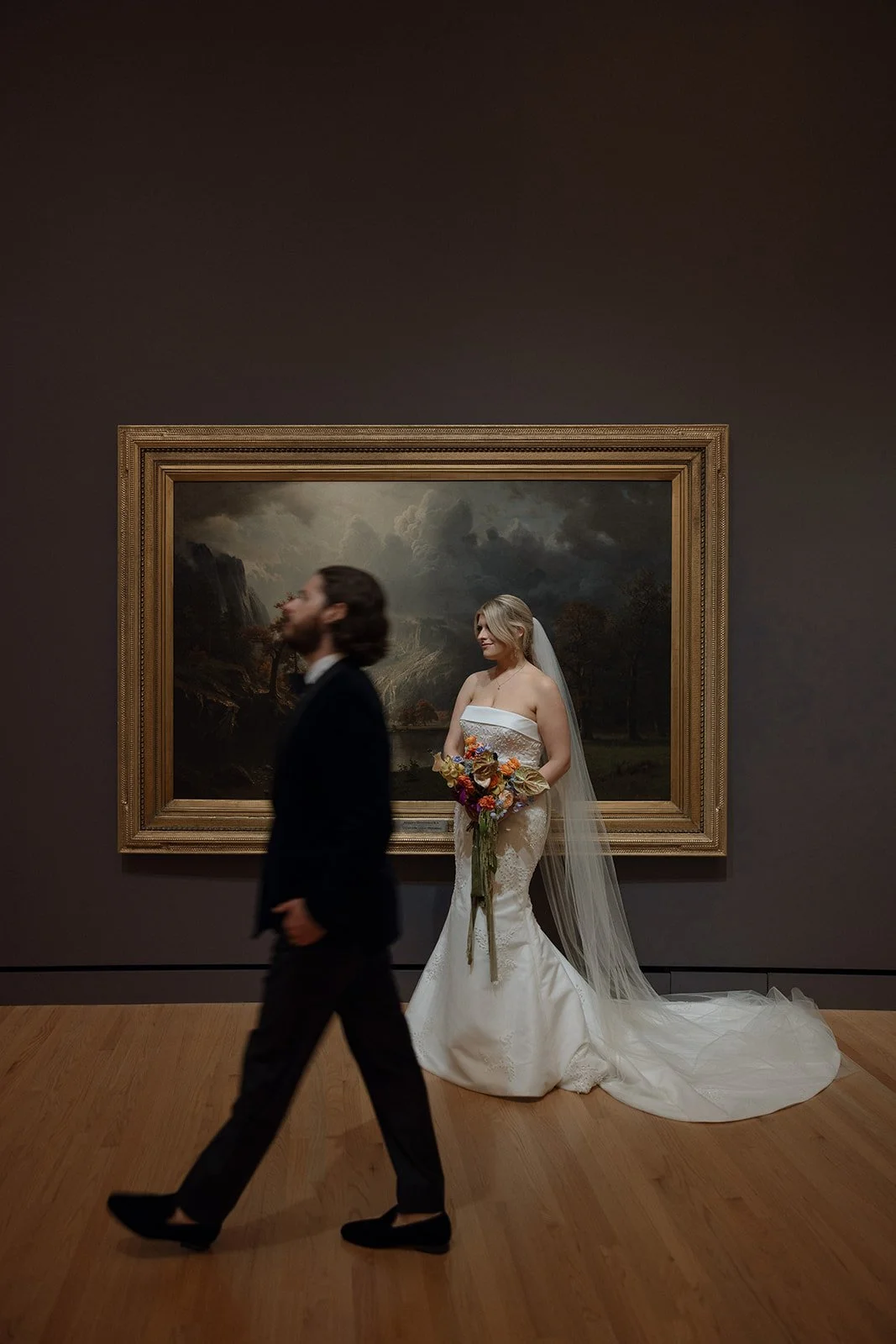 A bride in a wedding dress holding a colorful bouquet stands in front of a large framed painting in an art gallery. A man in a suit walks by in the foreground.