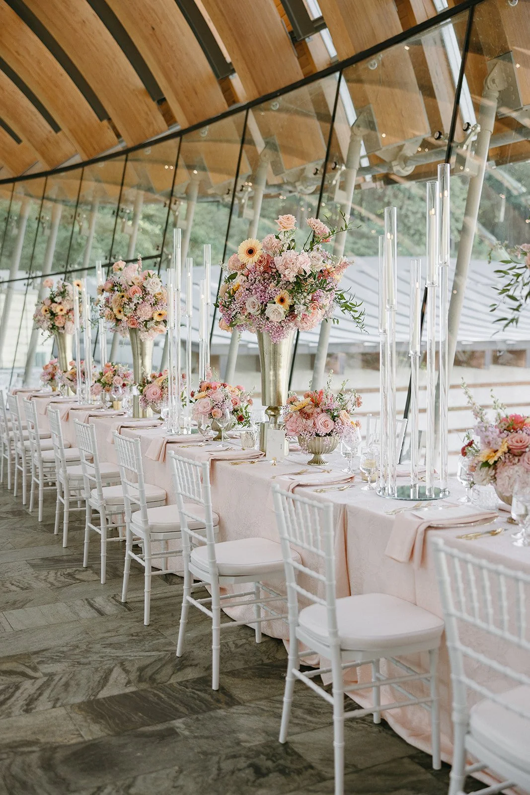 Elegant event table setting with floral centerpieces, tall glass candle holders, white chairs, and pink tablecloths in a venue with large windows and wooden ceiling.