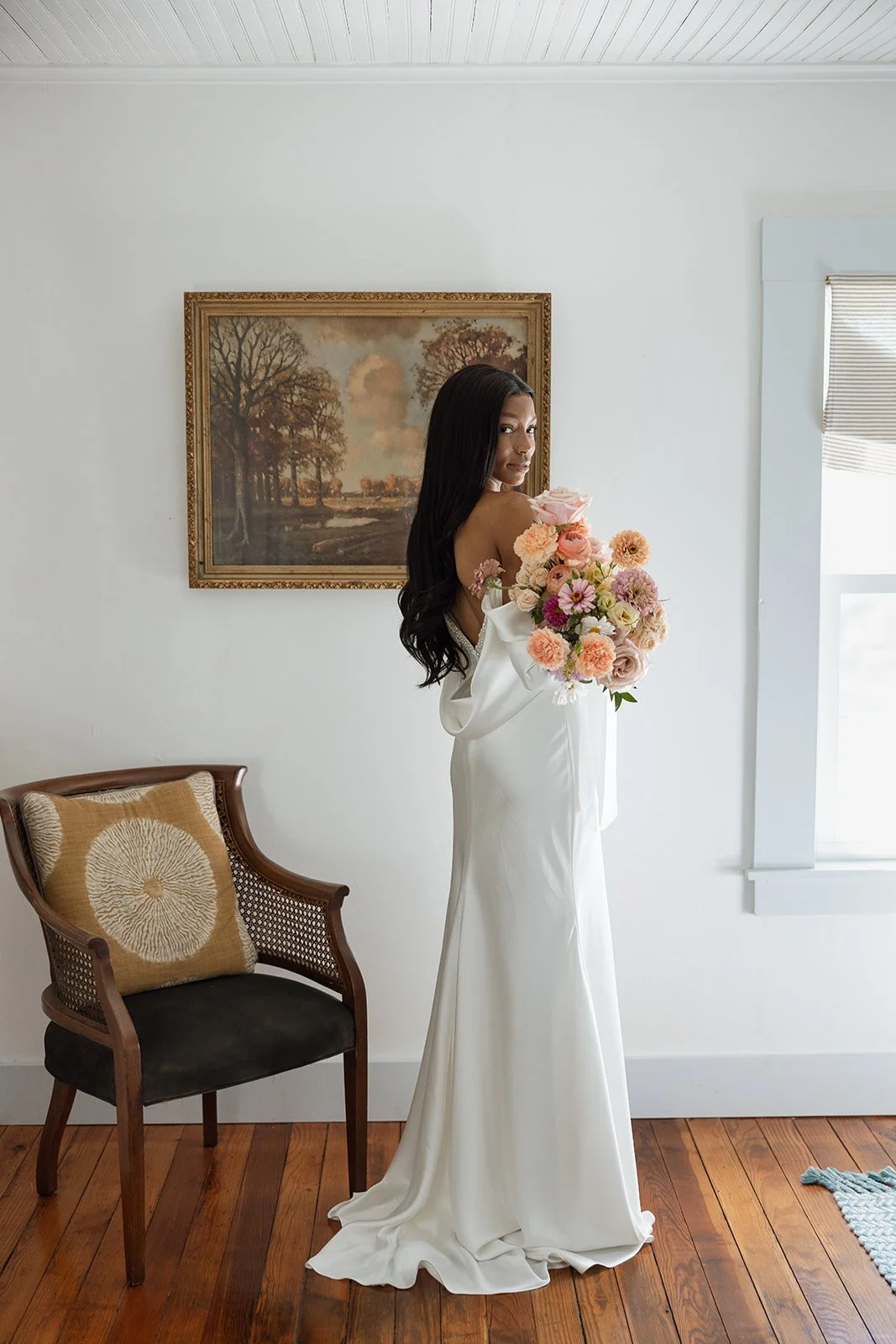 Bride in a white gown holding a bouquet, standing indoors next to a chair and art on the wall.