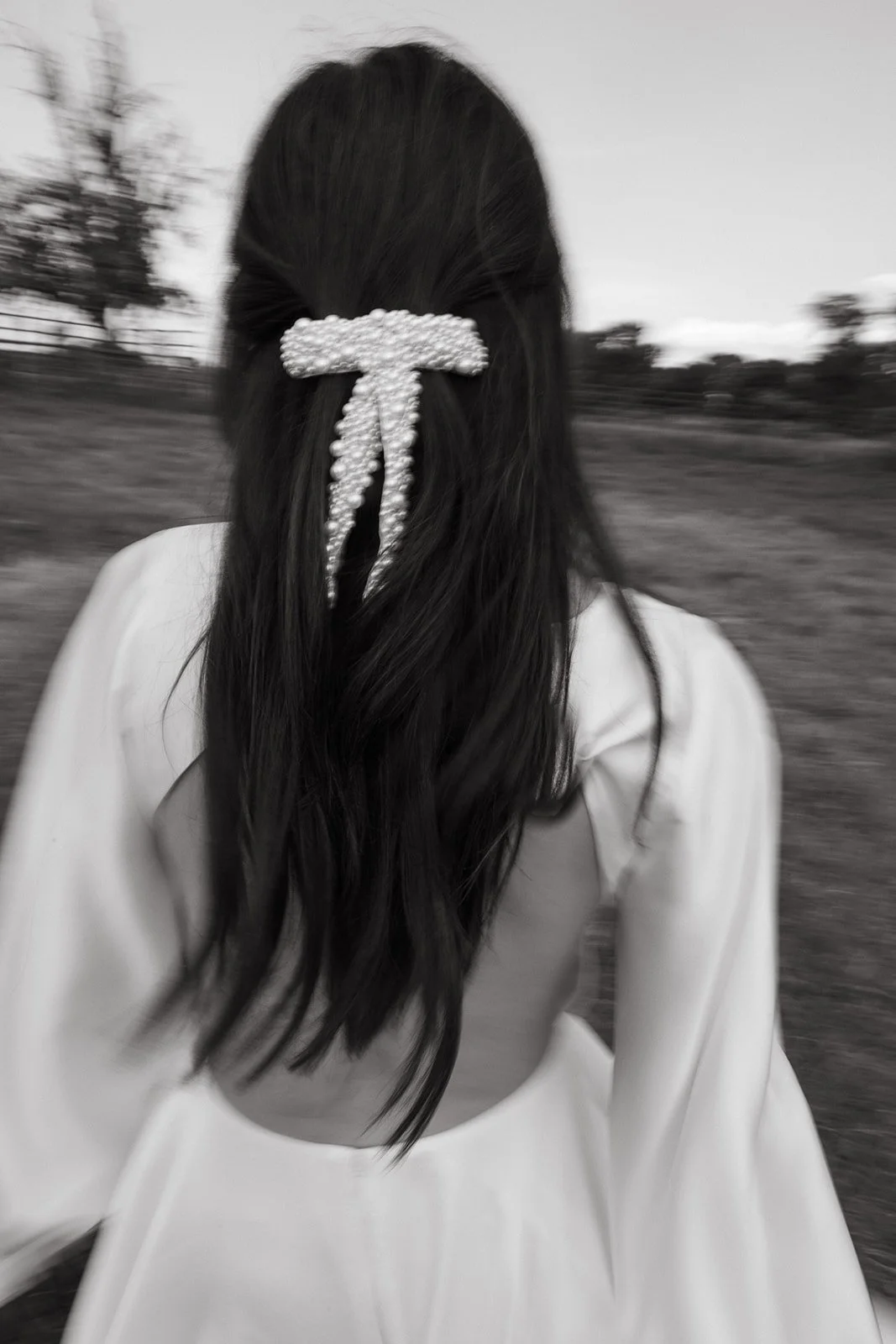 Black and white image of a person with long hair wearing a pearl hair accessory shaped like a bow, in an outdoor setting.