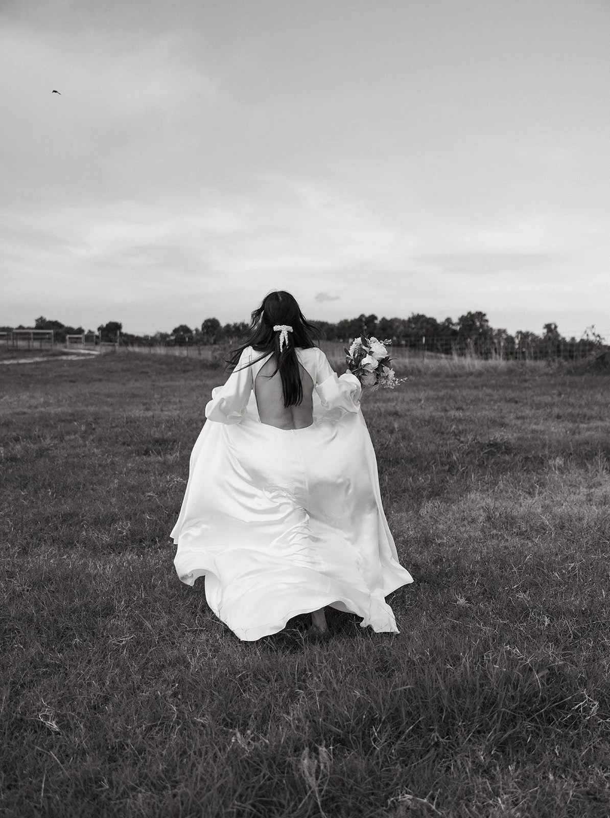 Bride in flowing white gown holding bouquet, walking through grassy field.