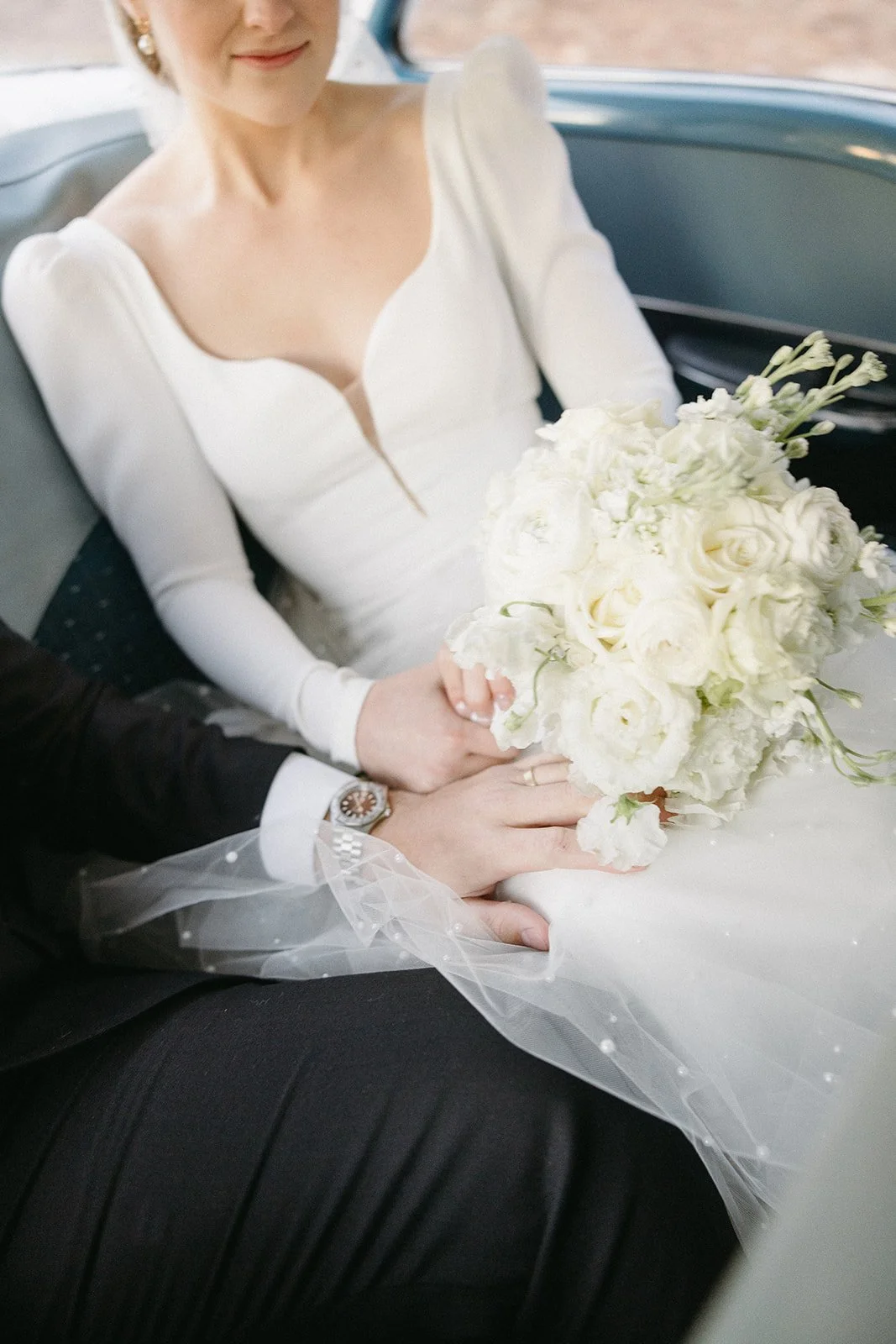 Bride in white dress holding bouquet of white flowers with groom's hand, sitting in car.