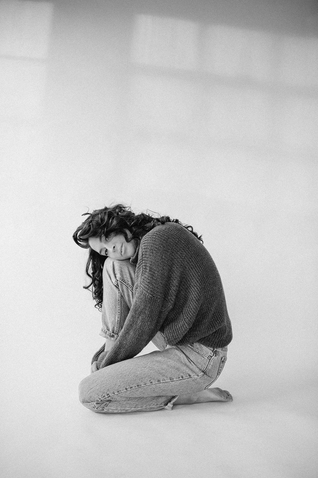 Black and white photo of a person sitting on the floor, wearing a sweater and jeans, with head resting on knees.