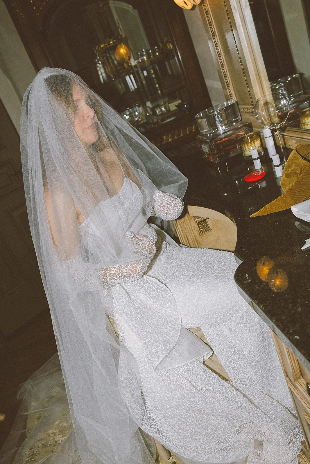 Bride in a lace wedding dress and veil sitting by a vanity with makeup items.