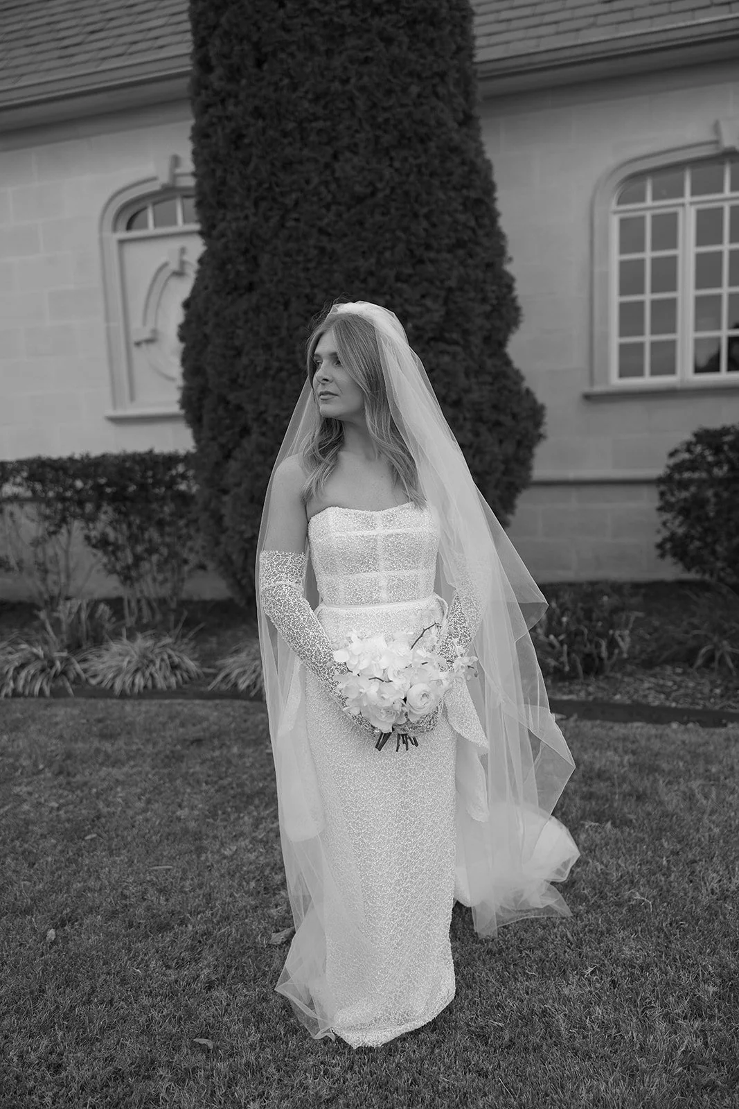 Bride standing outdoors in a wedding dress and veil, holding a bouquet.