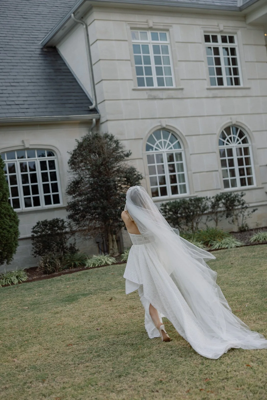 Bride walking towards a stone building with large windows, wearing a white wedding dress and a long veil, on a grassy lawn.