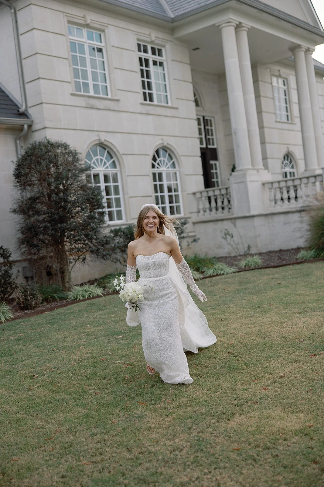 Bride in white gown holding bouquet, standing in front of a large building with arched windows and columns.