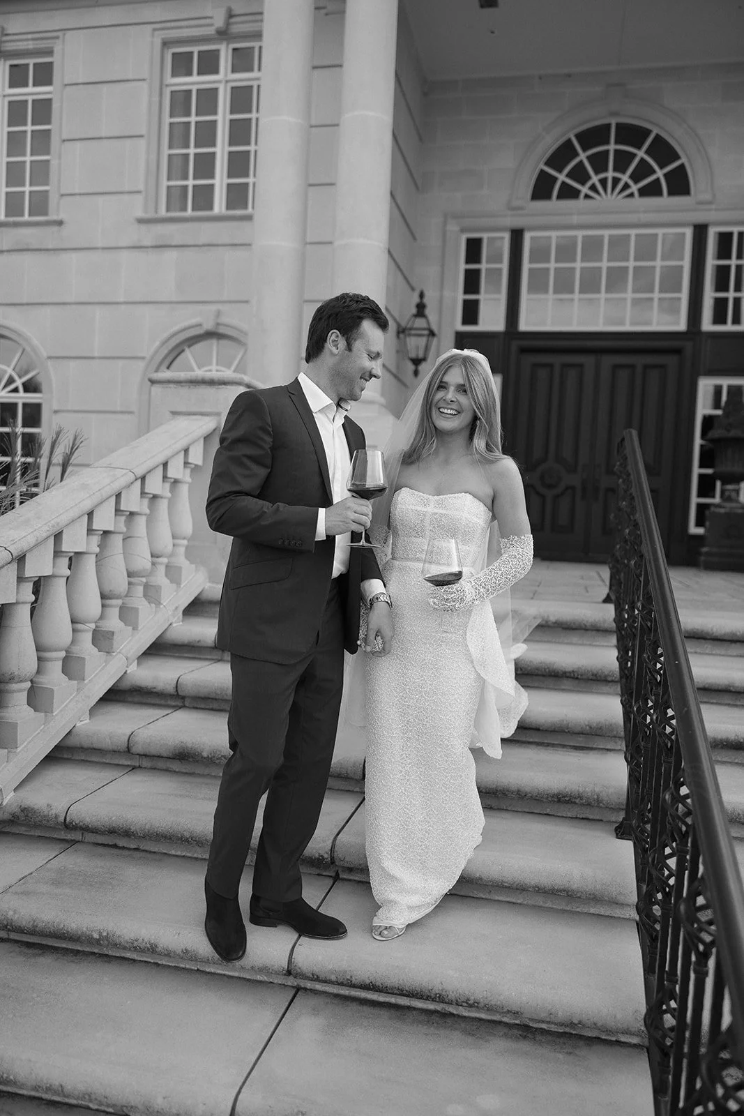 A couple dressed in formal attire, with the woman wearing a wedding gown and veil, hold glasses of wine on a staircase outside a building.