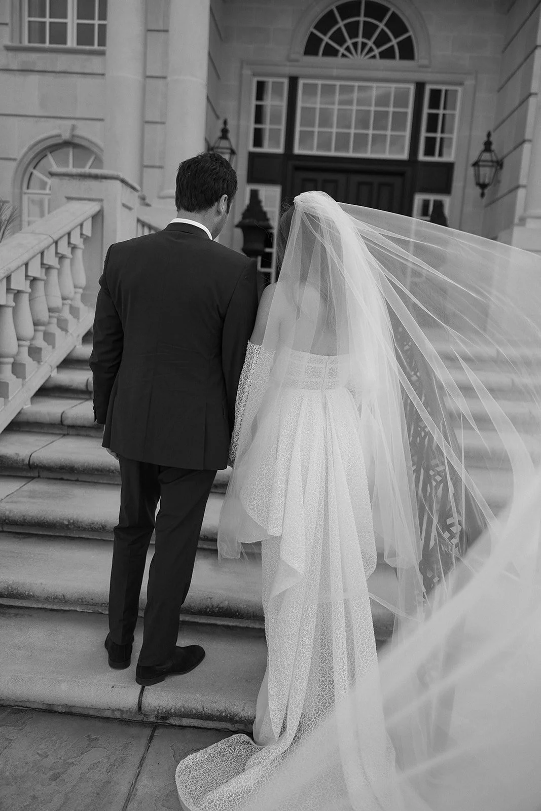 Bride and groom walking up steps to an elegant building, bride in a lace wedding dress and veil, groom in a suit, black and white.