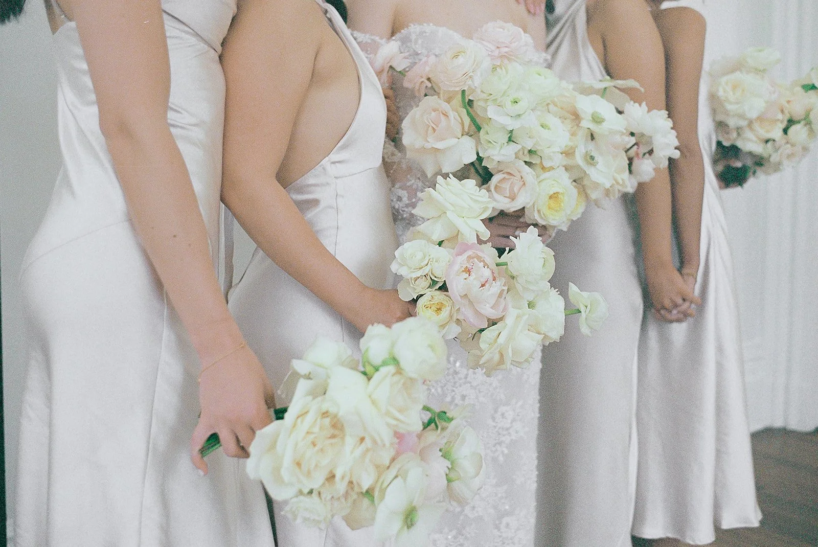 Group of women in light-colored dresses holding bouquets of white and blush flowers, likely at a wedding.