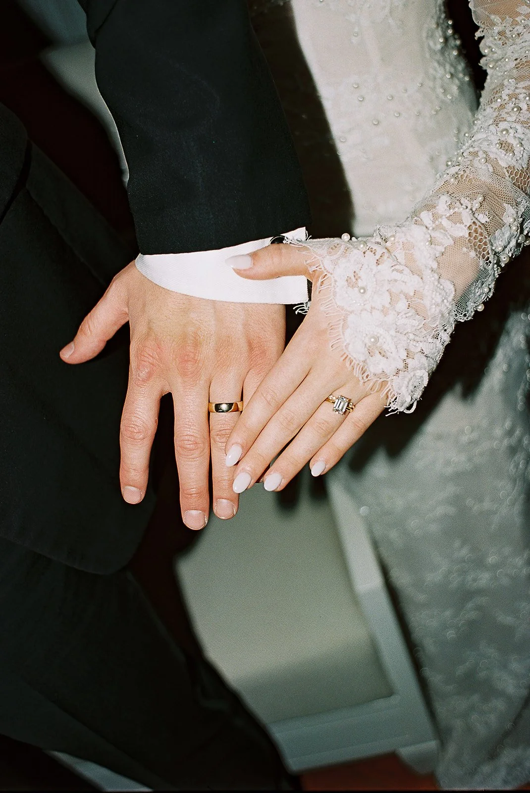 Close-up of a bride and groom's hands with wedding rings, showcasing the bride's lace wedding dress sleeve and manicured nails.