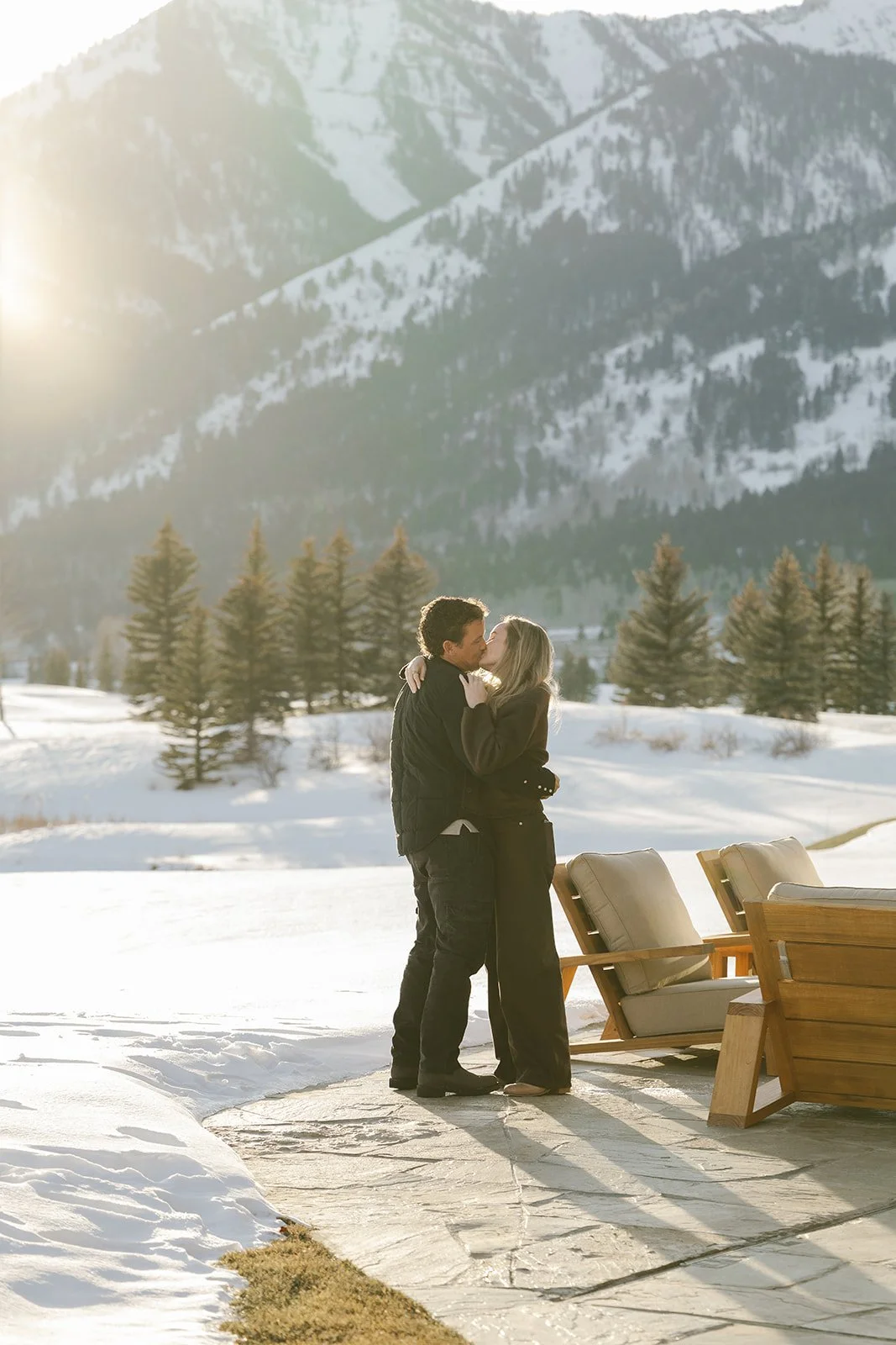 A couple kissing outdoors in a snowy landscape with mountains, evergreen trees, and outdoor furniture.