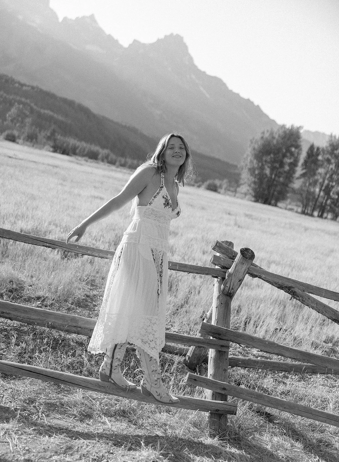A woman in a long lace dress and patterned boots standing on a wooden fence in a field with mountains in the background, smiling.