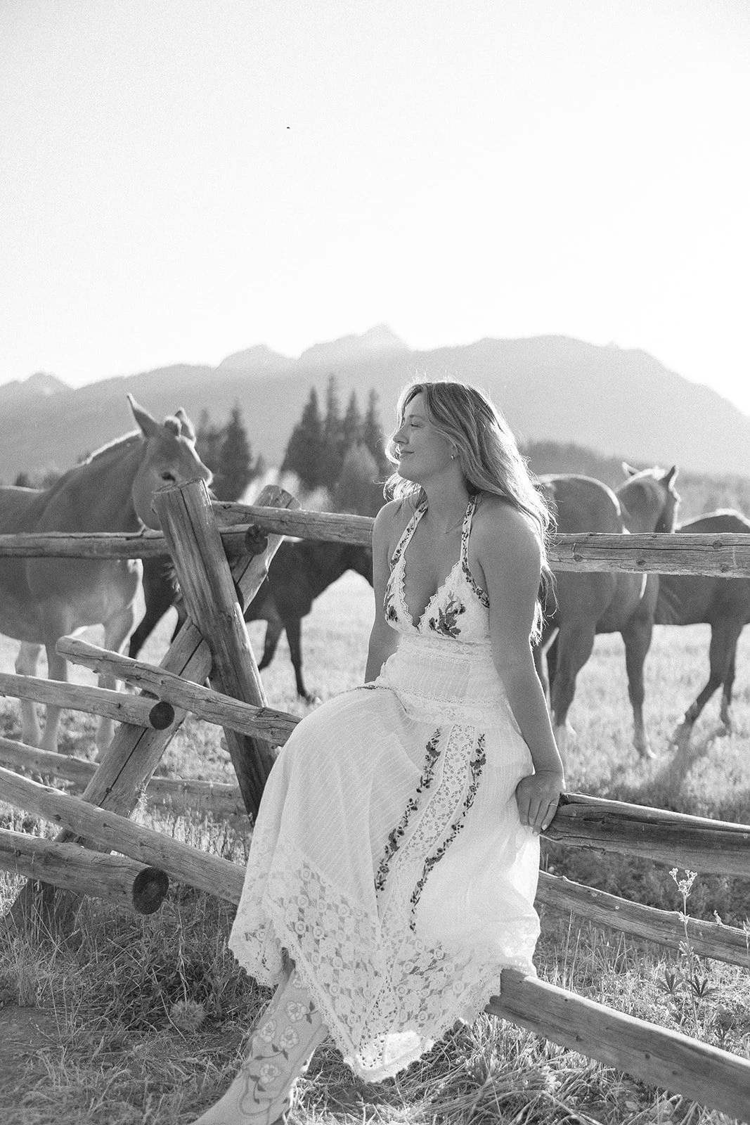 A woman in a white embroidered dress sits on a wooden fence in a rural landscape with horses and mountains in the background.