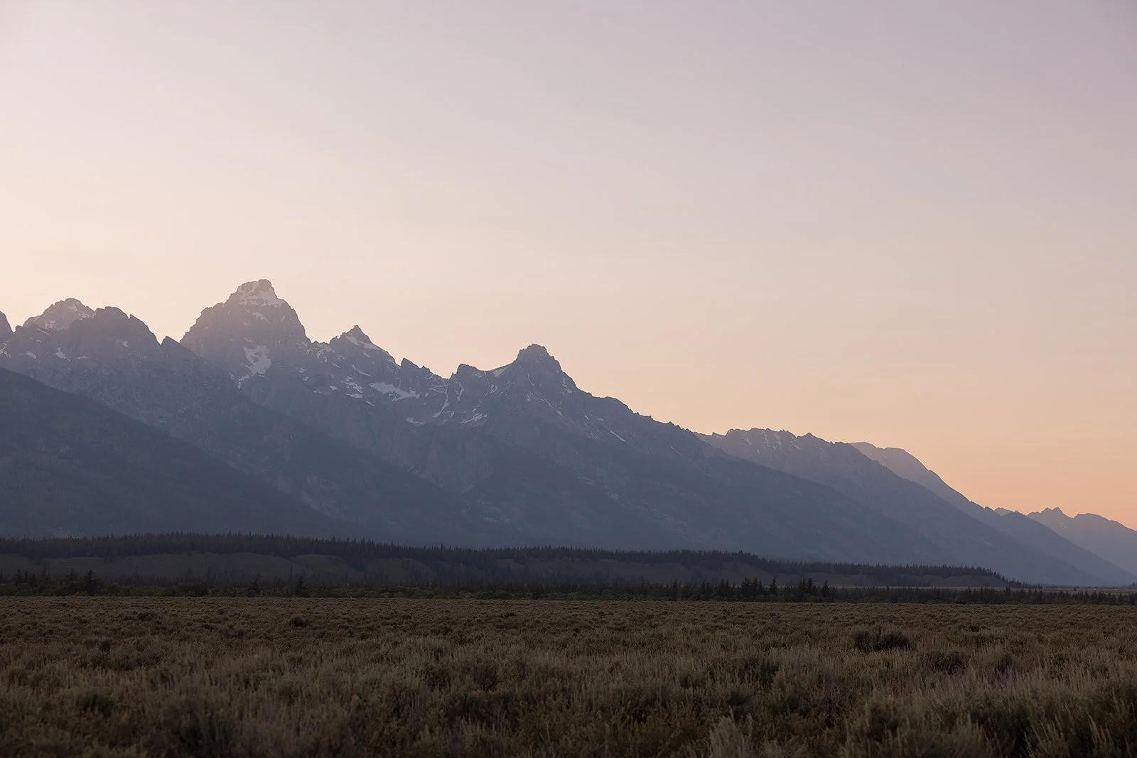 A mountain range at dusk with snow-capped peaks and a flat, grassy plain in the foreground.