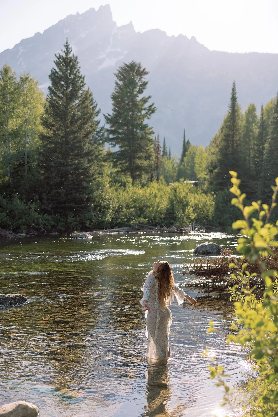 A woman with long hair in a white dress standing in a river, surrounded by lush green trees with a mountain in the background.