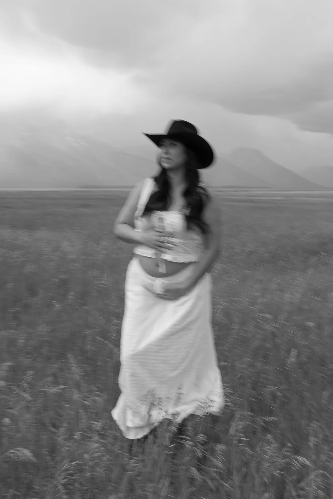 A woman wearing a wide-brimmed hat, sleeveless top, and long skirt standing in an open field with mountains and cloudy skies in the background.