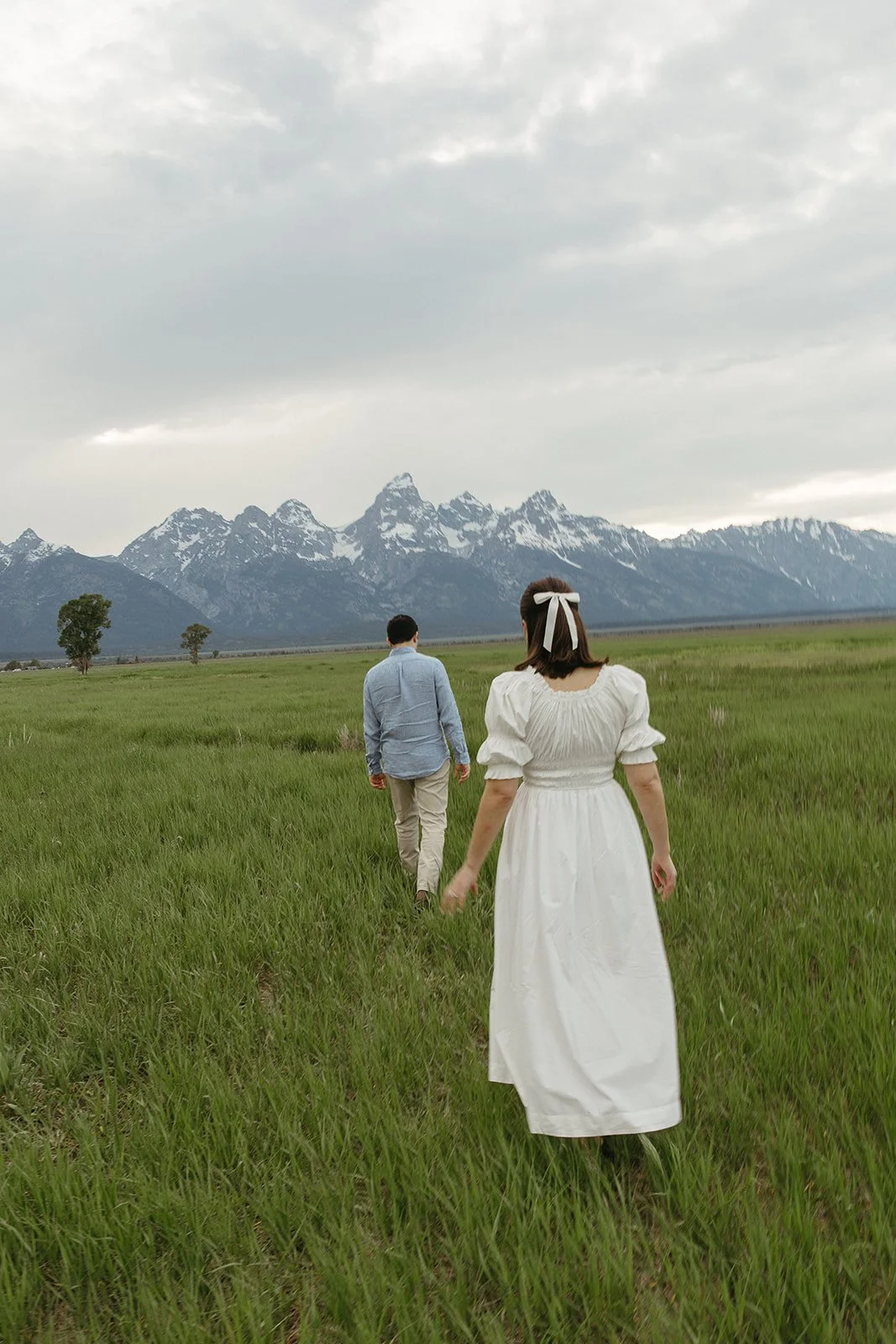 A woman in a white dress and a headband walking across a grassy field with a man in a blue shirt and beige pants, with snow-capped mountains in the background, under a cloudy sky.
