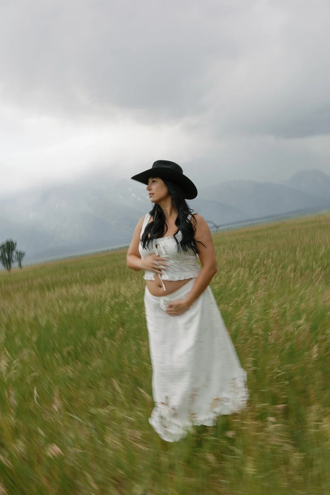 A woman with long black hair wearing a black cowboy hat, white crop top, and long white skirt standing in a grassy field with mountains and cloudy sky in the background.