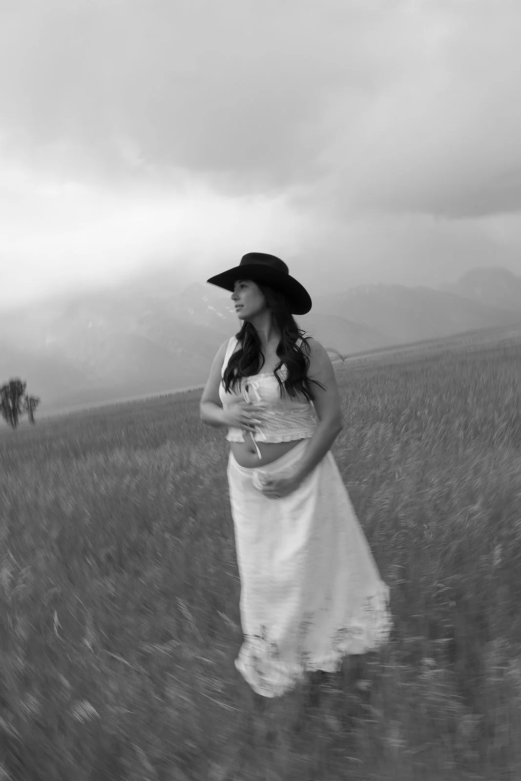 A woman in a long skirt and sleeveless top standing in a field with mountains and cloudy sky in the background, wearing a wide-brimmed hat.