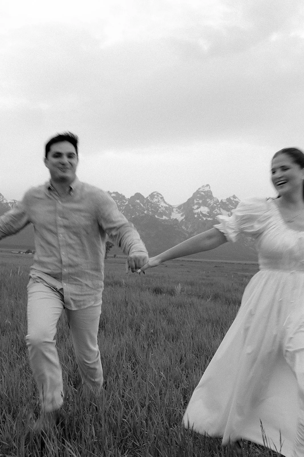 A happy couple holding hands and spinning in a grassy field with mountains in the background.