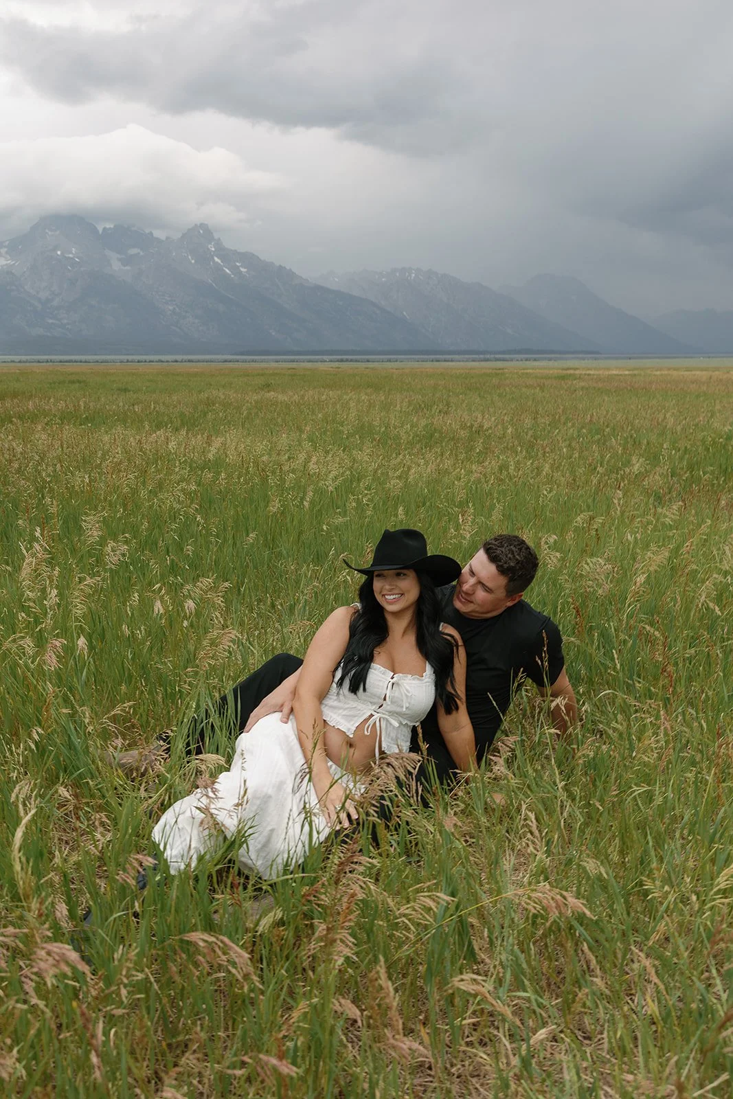 A couple sitting in a grassy field with mountains and cloudy sky in the background, smiling and enjoying the moment.