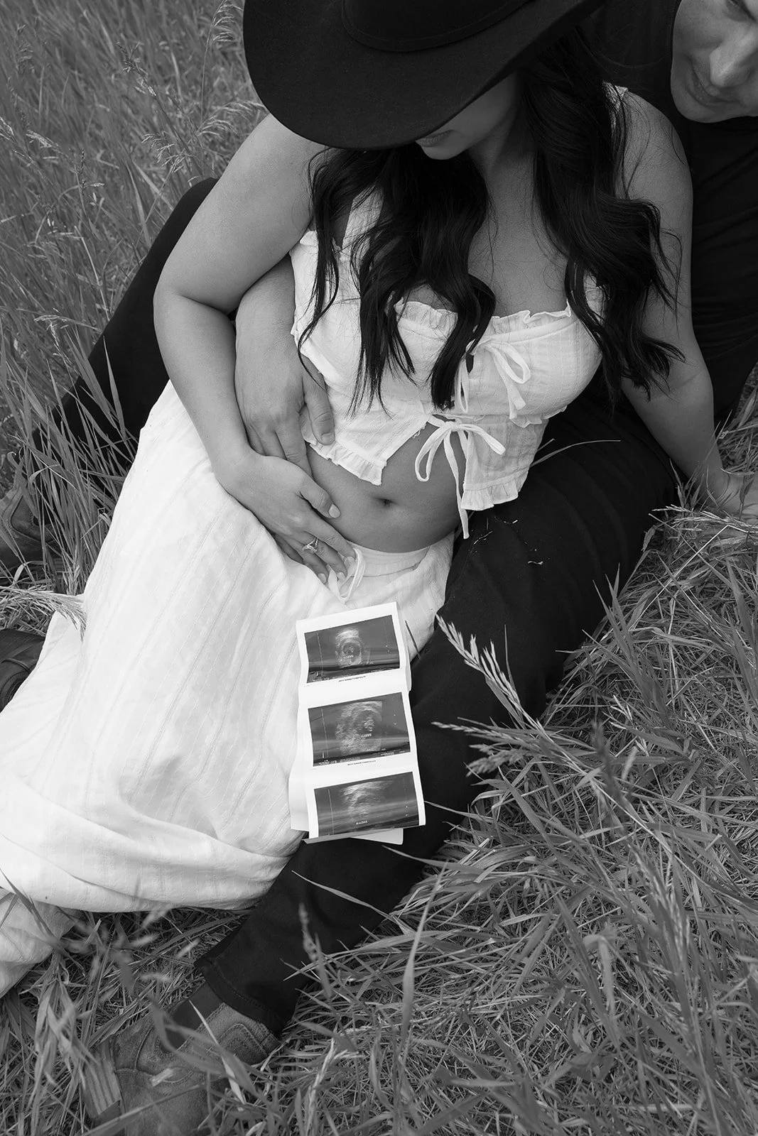 A woman in a white top and long skirt lying in grass, with a man behind her, both looking at ultrasound pictures on her lap.