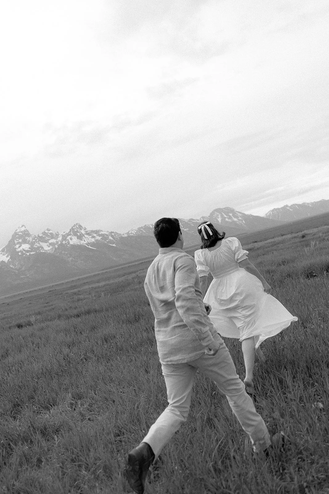 A man and woman in vintage clothing running through a grassy field with mountains in the background, captured in black and white.