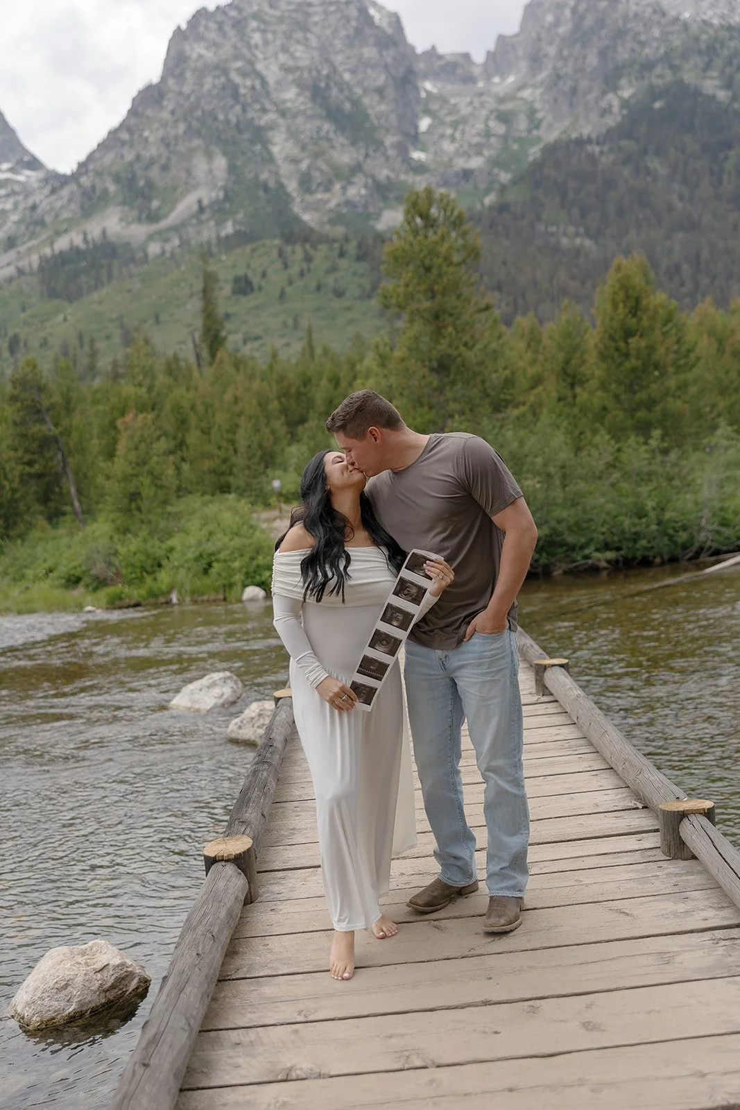 A couple sharing a kiss on a wooden bridge over a river with a backdrop of green trees and mountain peaks, and the woman holding ultrasound images.