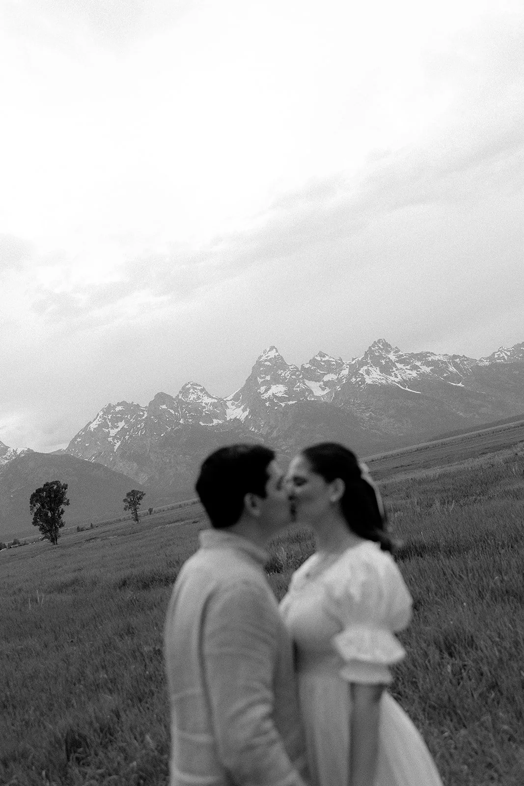 A black and white photo of a couple kissing in a grassy field with mountains in the background