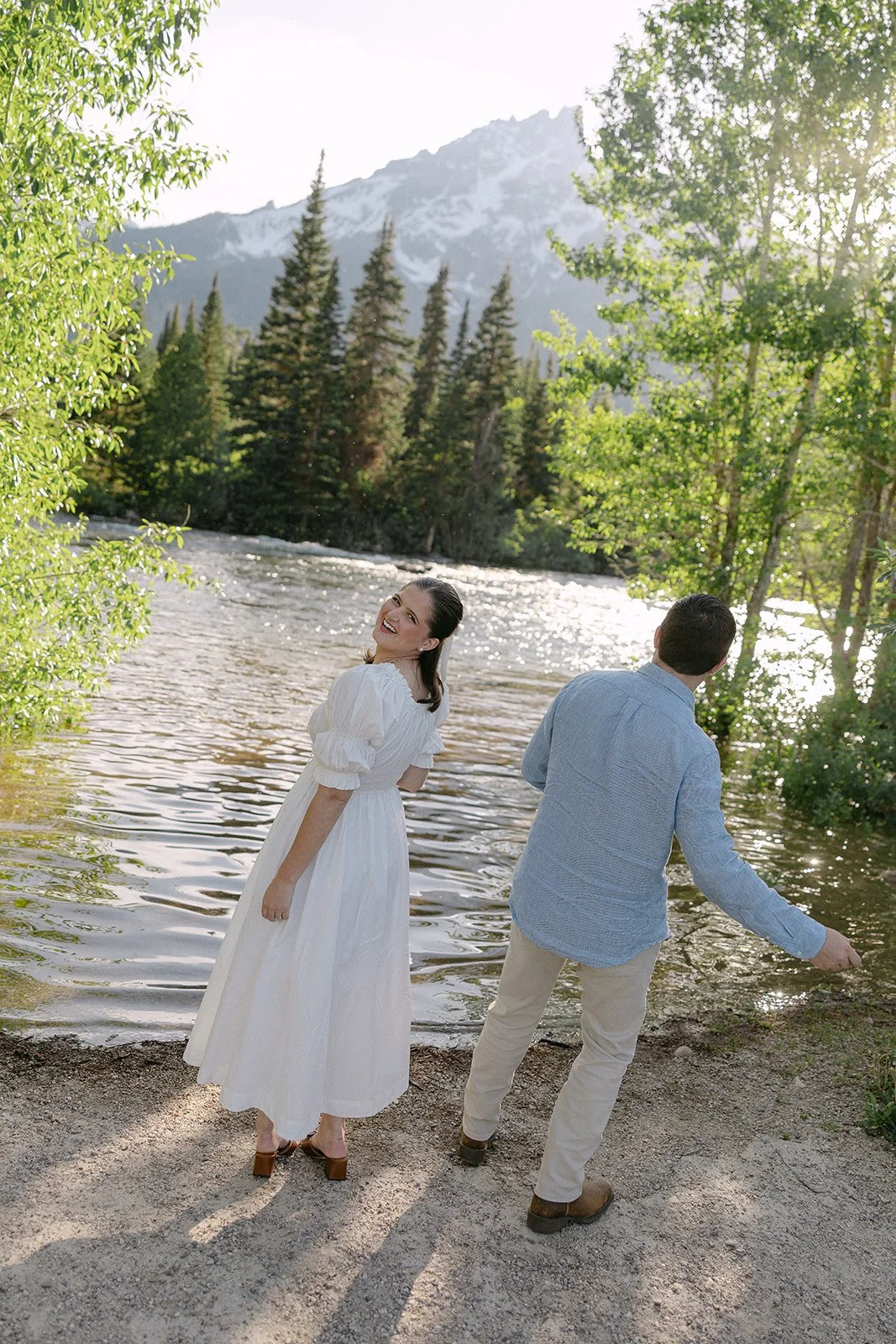 A woman in a white dress and a man in a light-colored shirt and pants on a riverbank in a forest with mountains in the background, as the woman smiles at the camera and the man looks away.