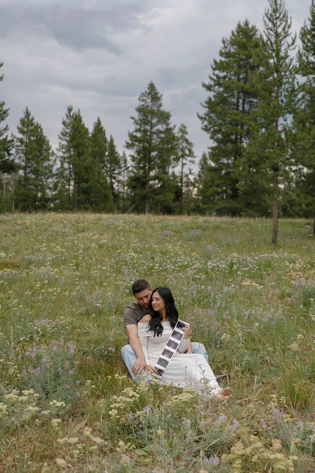 A couple sitting in a grassy field with wildflowers, holding ultrasound photos, embracing each other with a backdrop of pine trees and a cloudy sky.