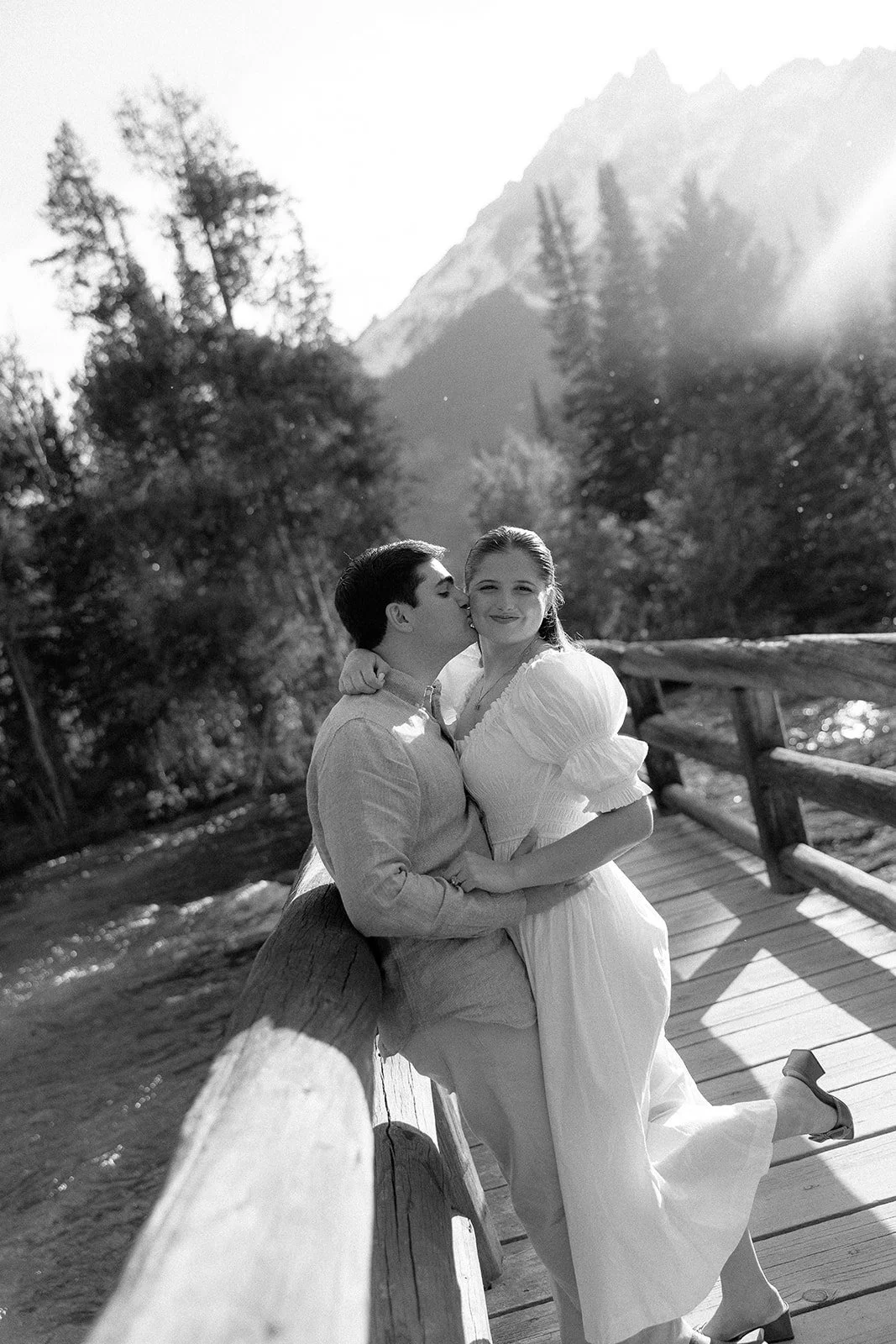 A black and white photo of a couple on a wooden bridge in a mountainous outdoor setting. The man is kissing the woman on her cheek while she smiles at the camera, wearing a white dress, and he is dressed in a light-colored shirt and pants.