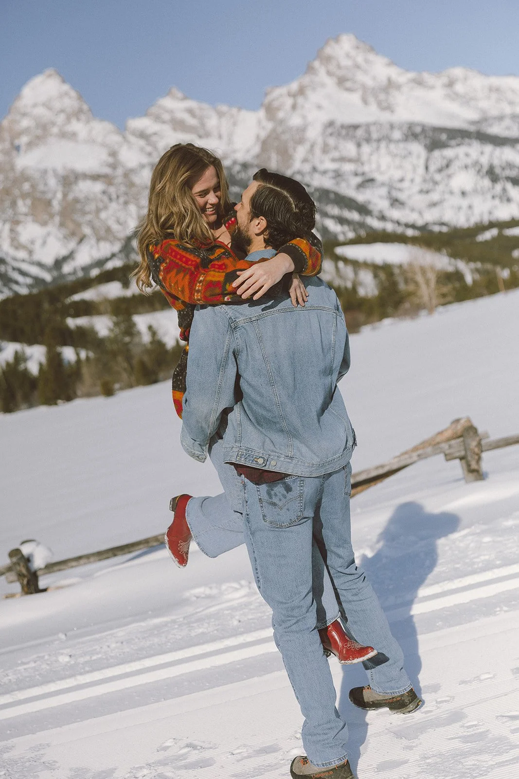 A couple in winter jackets and jeans outdoors in snow, with snowy mountains in the background, smiling and embracing.