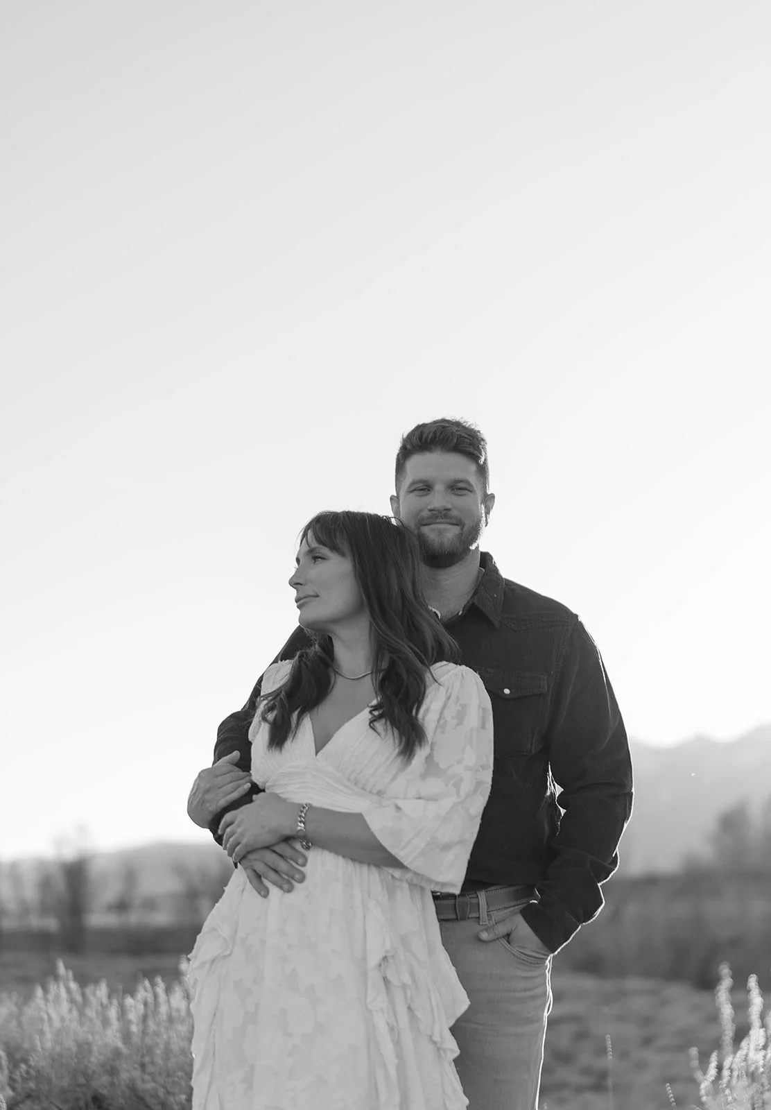 A black and white photo of a couple outdoors. The woman is in front, wearing a white dress, and the man stands behind her with his arm around her waist. The woman is looking to her left, and the man is looking at the camera with a slight smile.