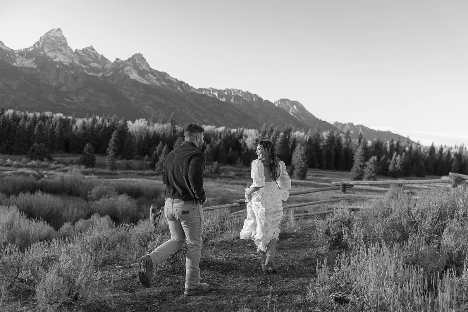 A man and woman walking along a dirt path through a mountainous landscape, with trees and bushes, in black and white.