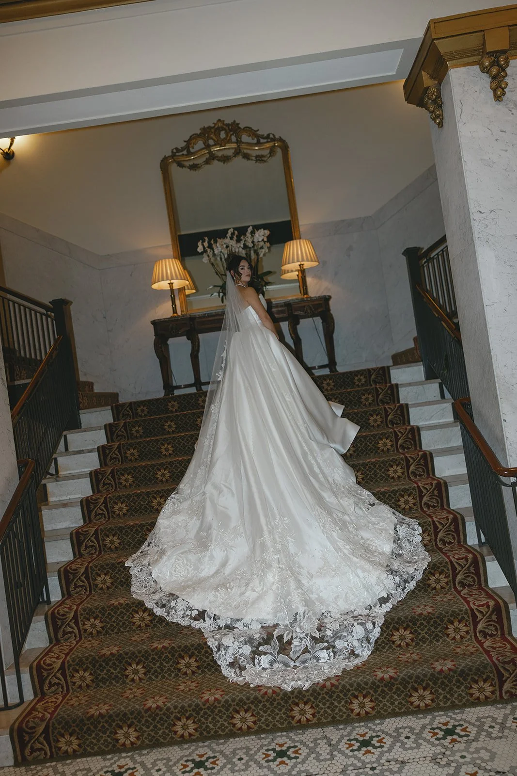 Bride in white gown on ornate staircase with mirror and lamps