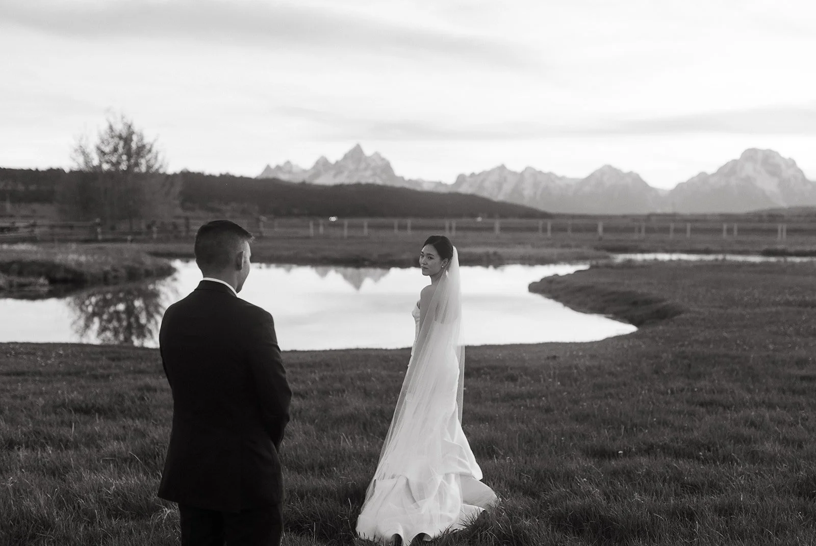 A black-and-white photograph of a bride and groom standing outdoors near a body of water with mountain ranges in the background. The bride is wearing a strapless wedding gown and veil, looking at the groom. The groom, in a suit, has his back turned t