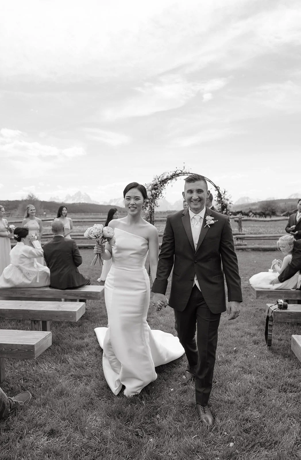 Black and white photo of a bride and groom walking hand-in-hand outdoors at a wedding ceremony. The bride is holding a bouquet and wearing a strapless wedding dress. The groom is in a dark suit with a boutonniere. In the background, seated guests and