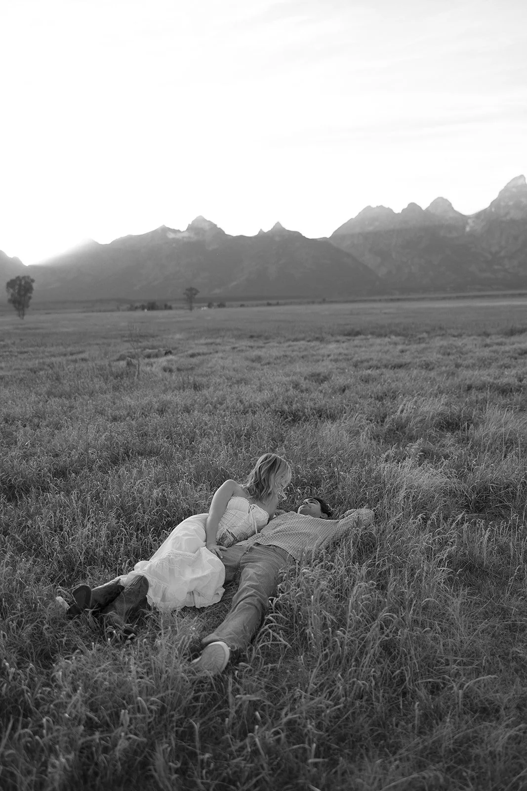 A woman and a man lying on a grassy field, the woman sitting beside the man who is reclining on the ground, set against a backdrop of mountain ranges in black and white.