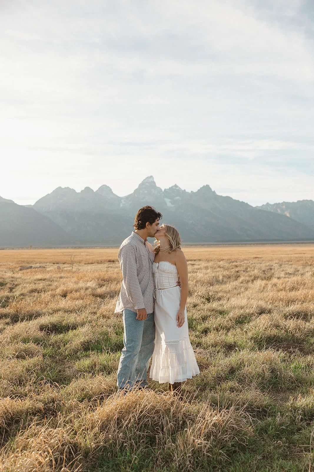 A couple sharing a kiss in a grassy field with mountains in the background, during a clear day.